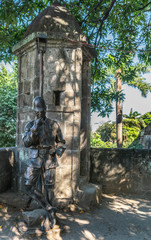 Manila, Philippines - March 5, 2019: Fort Santiago. Bronze statue of helmeted soldier leaning to rest against wall of cylindrical stone guard post under green foliage and some blue sky.