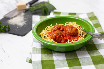 Spaghetti and Meatballs in Green Bowl on Green and White Checked Napkins on White Countertop; Grated Parmesan Cheese, Fresh Basil and Oregano on Black Cutting Board Beside