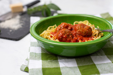 Spaghetti and Meatballs in Green Bowl on Green and White Checked Napkins on White Countertop; Grated Parmesan Cheese, Fresh Basil and Oregano on Black Cutting Board Beside