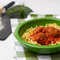 Spaghetti and Meatballs in Green Bowl on Green and White Checked Napkins on White Countertop; Grated Parmesan Cheese, Fresh Basil and Oregano on Black Cutting Board Beside