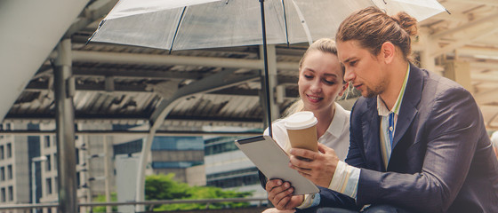 two caucasian business people holding umbrella sitting outdoor watching tablet computer with coffee...