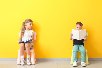 Little children with books sitting on chairs near color wall