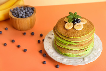 Tasty green pancakes with banana and blueberry on color background
