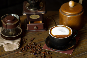 Closeup top view of hot cappucino cup with selective focus on coffee beans on wooden table with blurred grinder background