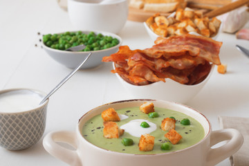 White wooden table with bowls of thick green pea soup embellished with roasted rusks, green peas, slices of grilled bacon, crushed black pepper and cream.
