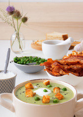 White wooden table with bowls of thick green pea soup embellished with roasted rusks, green peas, slices of grilled bacon, crushed black pepper and cream.
