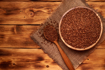 Uncooked buckwheat in a wooden bowl with the wooden spoon full of buckwheat, wooden background. Top view. Copy space in the left.