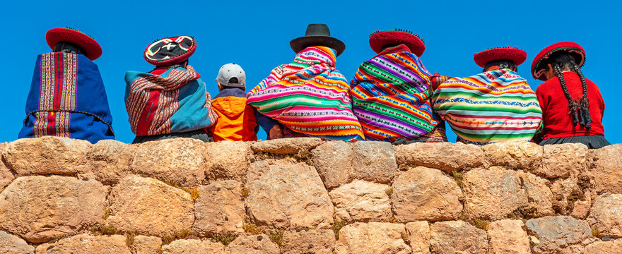 Panoramic photograph of Quechua indigenous women in traditional clothing with a boy sitting on an ancient Inca wall in Chinchero, Cusco Province, Peru.