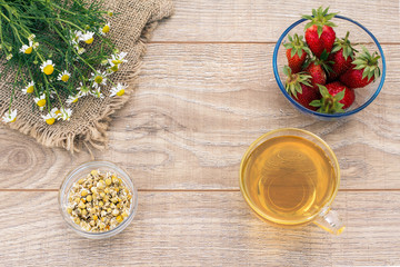Glass cup of green tea, stawberries with white chamomile flowers on wooden background.