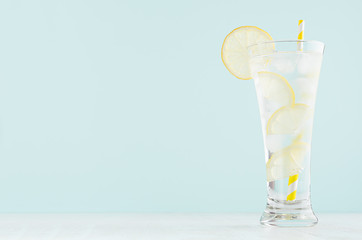 Infused fitness mineral tonic water with yellow lemon slices, ice cubes, yellow straw in elegant glass on white wood table, mint color wall.
