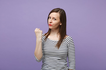 Strict angry young brunette woman in casual striped clothes posing isolated on violet purple background studio portrait. People lifestyle concept. Mock up copy space. Looking camera, Clenching fist.