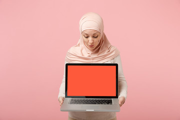 Young arabian muslim woman in hijab light clothes posing isolated on pink background. People religious Islam lifestyle concept. Mock up copy space. Holding laptop pc computer with blank empty screen.