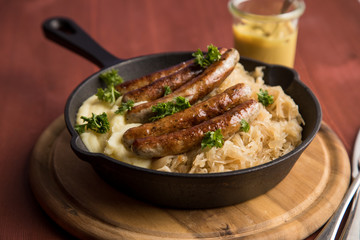 Kleine Bayrische Nürnberger Rostbratwürste mit Sauerkraut, Kartoffelpüree und Senf in rustikaler Gusseisenpfanne auf Holz Brett