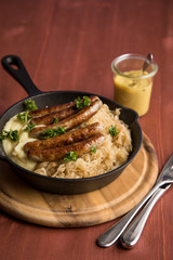 Kleine Bayrische Nürnberger Rostbratwürste mit Sauerkraut, Kartoffelpüree und Senf in rustikaler Gusseisenpfanne auf Holz Brett