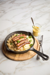 Kleine Bayrische Nürnberger Rostbratwürste mit Sauerkraut, Kartoffelpüree und Senf in rustikaler Gusseisenpfanne auf Holz Brett und Marmor Tisch