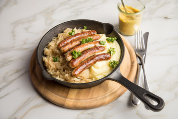 Kleine Bayrische Nürnberger Rostbratwürste mit Sauerkraut, Kartoffelpüree und Senf in rustikaler Gusseisenpfanne auf Holz Brett und Marmor Tisch