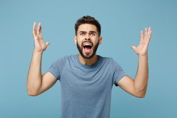 Naklejka premium Young angry shocked man in casual clothes posing isolated on blue background, studio portrait. People sincere emotions lifestyle concept. Mock up copy space. Looking up, screaming, spreading hands.