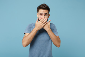 Fototapeta premium Young shocked concerned man in casual clothes posing isolated on blue wall background, studio portrait. People sincere emotions lifestyle concept. Mock up copy space. Covering mouth with hands.