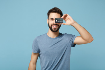 Young smiling handsome man in casual clothes posing isolated on blue wall background studio portrait. People sincere emotions lifestyle concept. Mock up copy space. Covering eye with credit bank card.