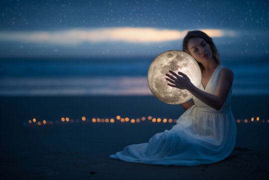 Tender image of a girl; female magic. Beautiful attractive girl on a night beach with sand and stars hugs the moon, art photo. On a dark background with space.