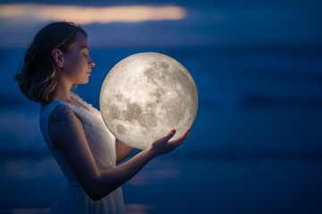 Naklejka premium Tender image of a girl. Female magic. Beautiful attractive girl on a night beach with sand hugs the moon, art photo. On a dark background with space. Riddle, astrology, zodiac. Banner.