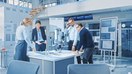 Diverse Team of Industrial Robotics Engineers Gathered Around Table With Robot Arm, They Manipulate and Program it to Pick Up and Move Metal Component. Bright Facility, Creative People