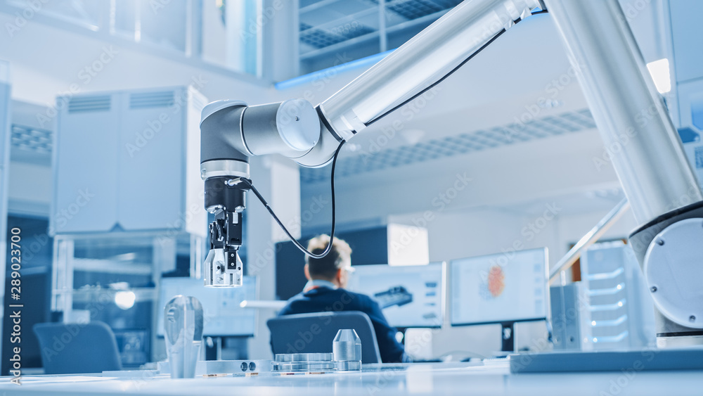 Leinwandbilder Modern Robot Arm Picks up, Lifts and Moves Metal Component as Programmed. In the Background Industrial Robotics Engineers Working on Software, Programming and Machine Learning in Engineering Facility