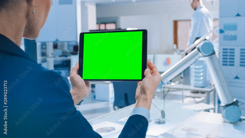 Poster Engineer Holds Green Mock-up Screen Digital Tablet Computer. In the Background Team of Industrial Engineers Working with Functional Robot Arm in Modern Bright Research Facility
