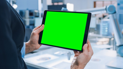 Engineer Holds Green Mock-up Screen Digital Tablet Computer. In the Background Team of Industrial Engineers Working with Functional Robot Arm in Modern Bright Research Facility