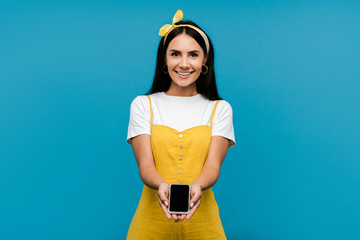 happy woman in yellow dress holding smartphone with blank screen isolated on blue