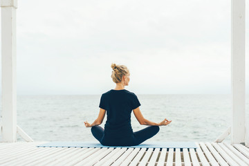 A woman in sportswear is sitting in a lotus position with her back to the camera and meditating while looking at the sea. Calm peaceful mood. Mental comfort.