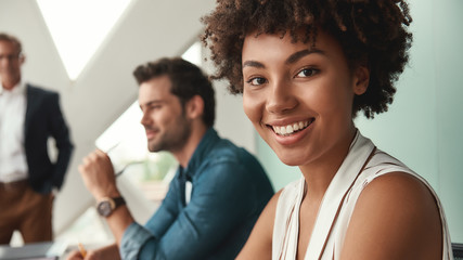 Fototapeta premium Feeling happy. Young afro american woman is looking at camera and smiling while working with colleagues together at office