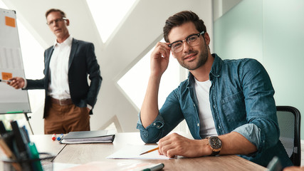 Smart and confident. Young and handsome man adjusting eyeglasses and looking at camera with smile while his boss standing near whiteboard and explaining something on the background