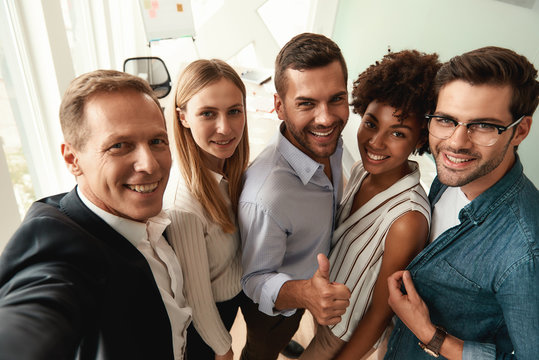 Group of cheerful colleagues taking selfie and gesturing while standing in the modern office