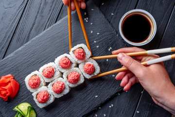 Hand with chopsticks takes sushi from Seth sushi rolls with cream cheese, rice and salmon on a black board decorated with ginger and wassabi on a dark wooden background. Japanese food