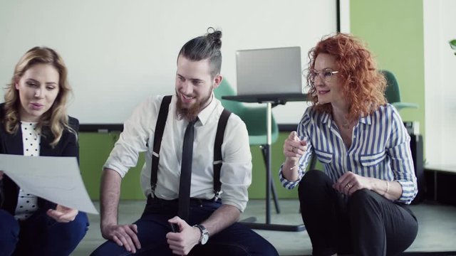 A group of young business people sitting on the floor in an office, talking.