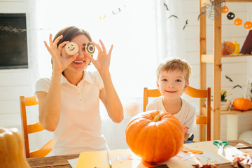 Fototapeta premium Happy Halloween. Young woman with her little son are preparing to Halloween on kitchen. Mother with son are having fun with pumpkins. Happy Halloween concept