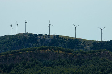 Six wind turbines on top of a forested mountain in a haze against a pale sky (Rhodes, Greece)