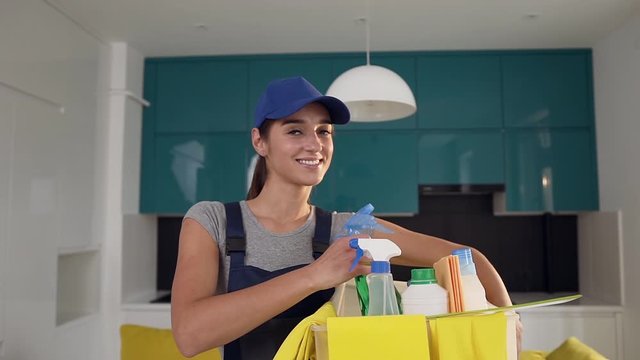 Cheerful cleaning service woman holding the box with special detergents and spraying item for cleaning,standing in the cuisine