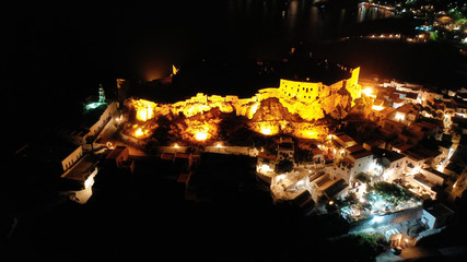 Aerial drone night shot of iconic illuminated medieval fortified castle overlooking the deep blue Aegean sea in Chora of Astypalaia, Dodecanese islands, Greece