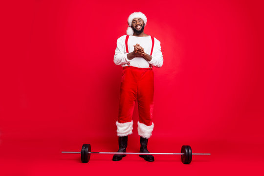 Full body photo of cheerful bearded sportsman black santa claus in xmas cap ready to carry barbell wearing white sweater pants trousers isolated over red background