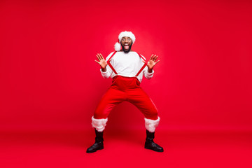 Fototapeta na wymiar Full body photo of cheerful hipster afro american santa touching his suspenders screaming wearing trousers pants white pullover isolated over red background