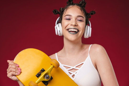 Image of happy punk girl with bizarre hairstyle and dark lipstick holding skateboard while listening to music with headphones