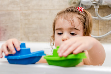 Happy little baby girl sitting in bath tub playing with colorful boats toys in the bathroom.