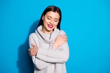 Portrait of positive woman cuddling herself with eyes closed wearing grey woolen pullover isolated over blue background