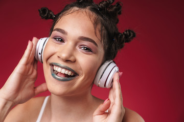 Fototapeta premium Image of positive punk girl with bizarre hairstyle and dark lipstick smiling while listening to music with headphones