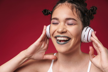 Fototapeta premium Image of excited punk girl with bizarre hairstyle and dark lipstick smiling while listening to music with headphones