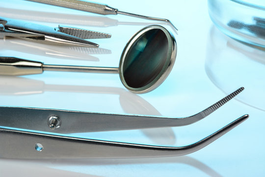 Dentist equipment: dental mirror, explorer, kornzange, forceps, dental burs in Petri dish. Dental tools on blue glossy surface, closeup.