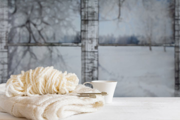 Cozy winter still life: mug of hot tea and warm woolen knitting on vintage windowsill against snow landscape from outside.