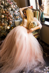 young blonde glamour girl in pink air dress is sitting like princess in the armchair with hand near head, looking away and smiling cute near window and christmas tree. christmas concept